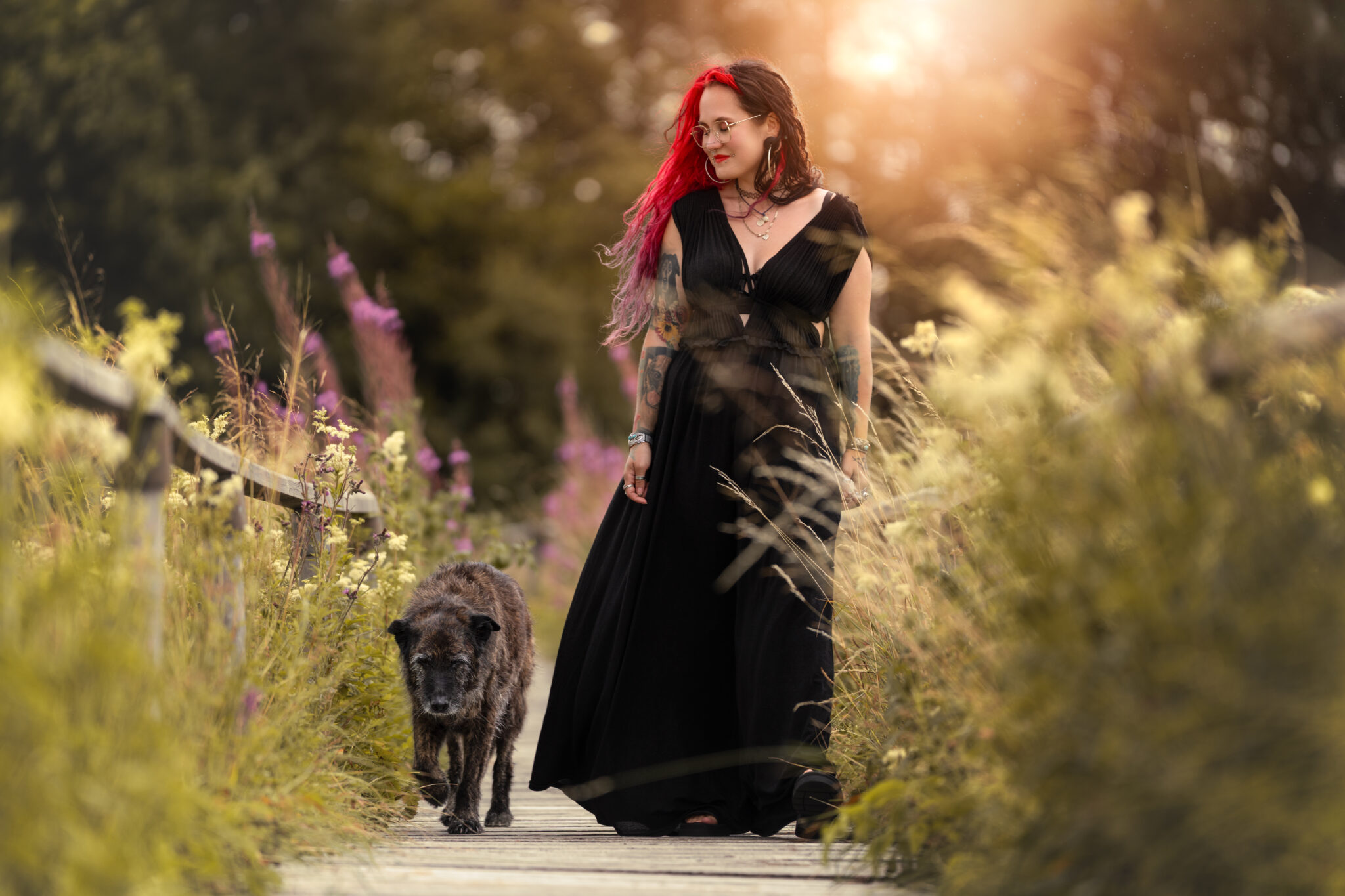 Tierfotograf Tiershooting: Frau mit Hund auf Holzweg im Sonnenuntergang, Wildblumen überall.