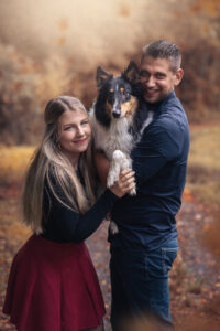 Lächelndes Paar stolz beim Tiershooting mit Hund, Herbstblätter im Hintergrund.